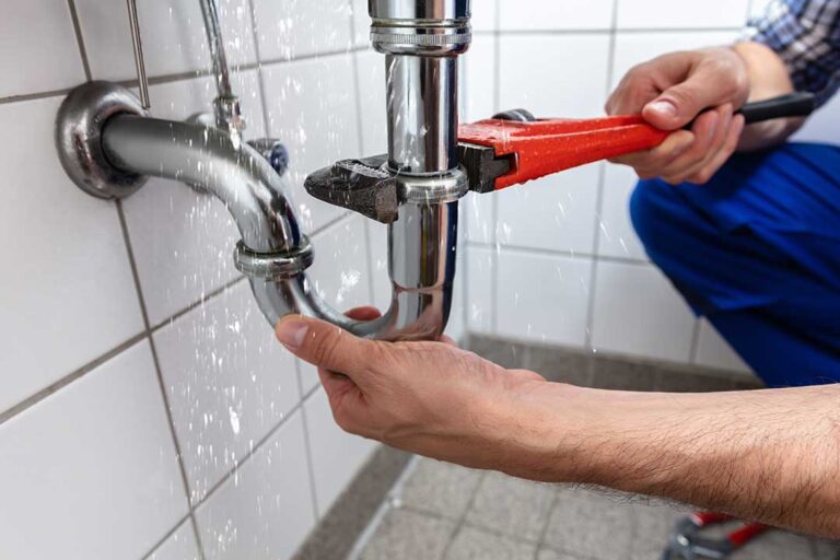 Homeowner inspecting aging pipes beneath a kitchen sink with a flashlight and small bucket