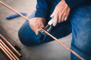 Homeowner examining a dripping faucet beneath a kitchen sink with a bucket and adjustable wrench nearby.